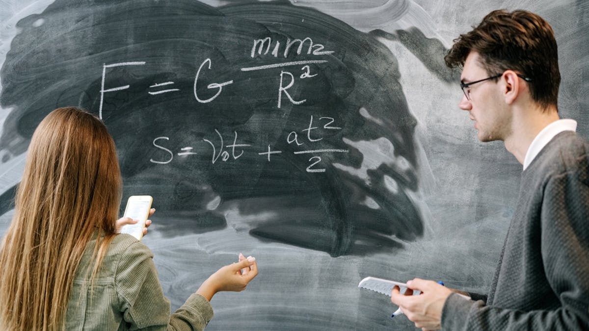 Two students discussing physics equations on a chalkboard in a classroom setting.